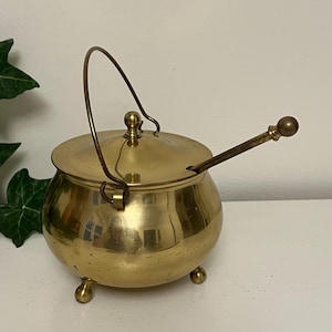 May include: A small, round brass pot with a lid, handle, and three small feet. A brass spoon rests in the pot. The pot has a shiny, reflective surface. The handle is arched, and the lid has a small knob.