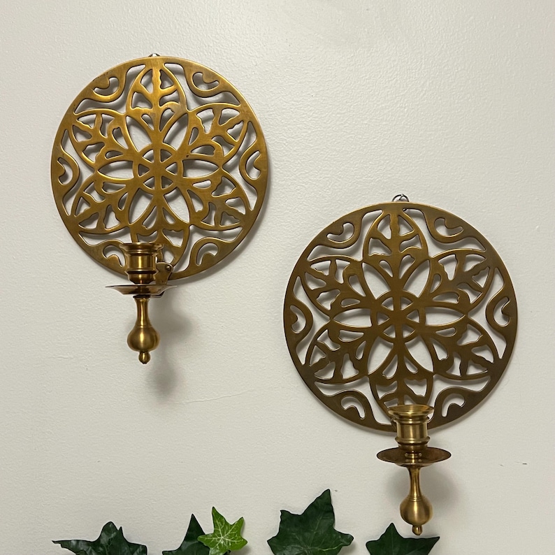 May include: Two gold-colored, circular wall sconces with intricate, openwork floral designs. Each sconce features a candle holder. The sconces are mounted on a white wall, creating a decorative and elegant display.