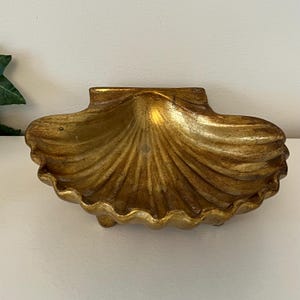 May include: A decorative, gold-colored seashell-shaped dish. The dish has a textured surface and a scalloped edge. The interior of the shell is smooth and shiny. The dish is resting on a white surface.