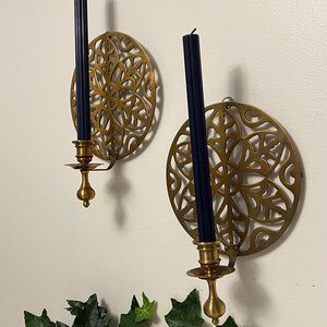 May include: Two brass wall sconces with navy blue candles. Each sconce features a circular, openwork design. The candle holders are attached to the wall with a small hook. Green leaves are visible at the bottom of the image.