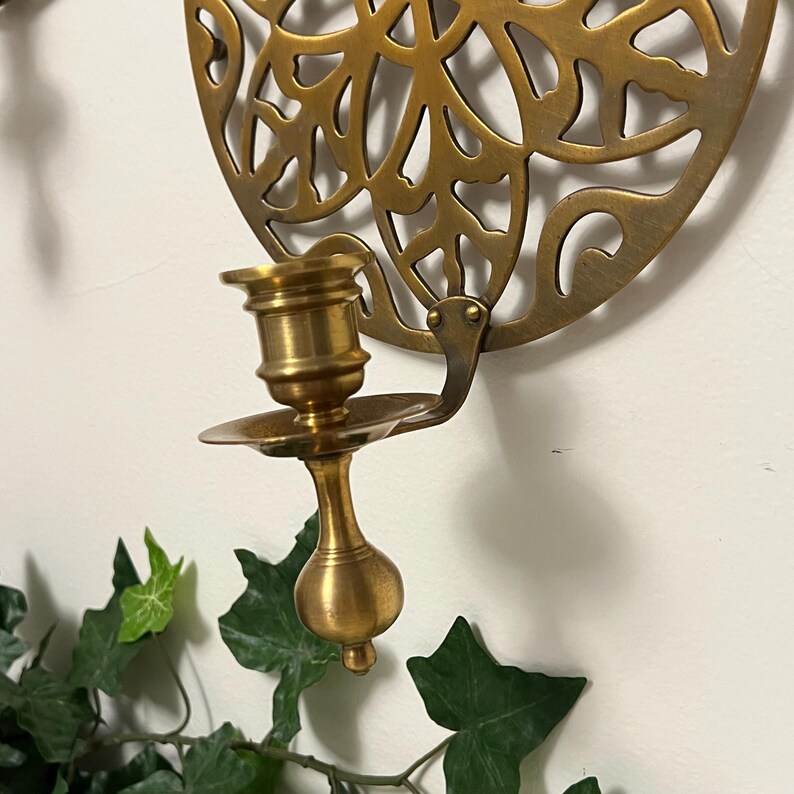 May include: A brass wall-mounted candle holder with a decorative, openwork design. The candle holder has a cup-shaped base and a small, round dish to catch wax. The background is a white wall with green ivy.