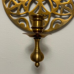 May include: A brass wall-mounted candle holder with a decorative, openwork design. The candle holder has a cup-shaped base and a small, round dish to catch wax. The overall design is ornate and vintage.