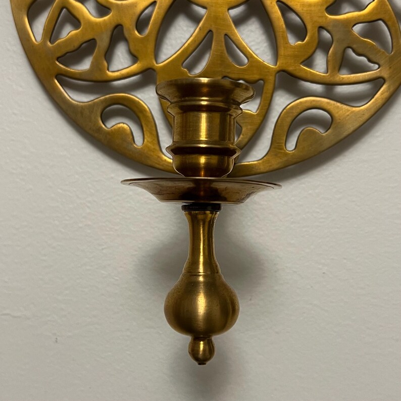 May include: A brass wall-mounted candle holder with a decorative, openwork design. The candle holder has a cup-shaped base and a small, round dish to catch wax. The overall design is ornate and vintage.