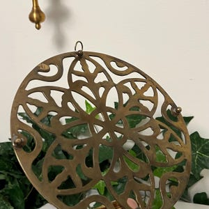 May include: A round, antique brass trivet with a decorative openwork design. The trivet features a floral pattern and small, rounded feet. A brass hook is attached to the top for hanging. The background includes green foliage and a white wall.