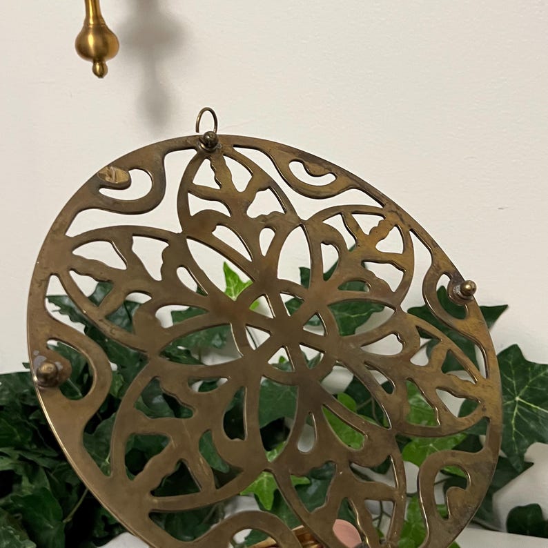 May include: A round, antique brass trivet with a decorative openwork design. The trivet features a floral pattern and small, rounded feet. A brass hook is attached to the top for hanging. The background includes green foliage and a white wall.