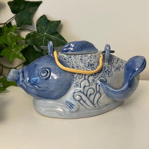 May include: A blue and white ceramic teapot shaped like a fish. The teapot has a lid, a spout, and a handle. The fish-shaped teapot is decorated with floral patterns and a yellow handle.