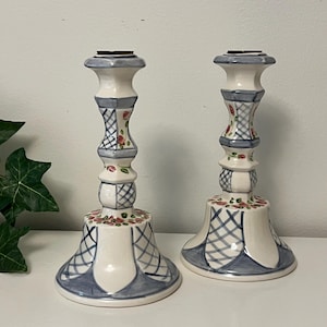 May include: Pair of ceramic candlesticks. The candlesticks are white with blue and red floral and lattice designs. The base is flared, and the top has a small, flat surface for holding a candle. The candlesticks are decorative.