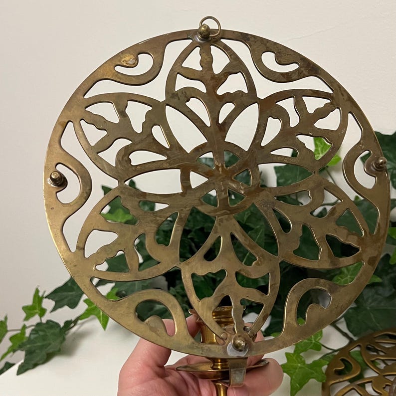 May include: A vintage brass candle holder with a circular, openwork design. The intricate pattern features floral and leaf motifs. The candle holder has a small loop at the top for hanging and a base for the candle. The brass has a warm, aged patina.