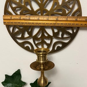 May include: A brass candle holder with a decorative, circular filigree design. The candle holder is approximately 9 inches in diameter. A wooden ruler is placed across the top of the candle holder to measure its size.