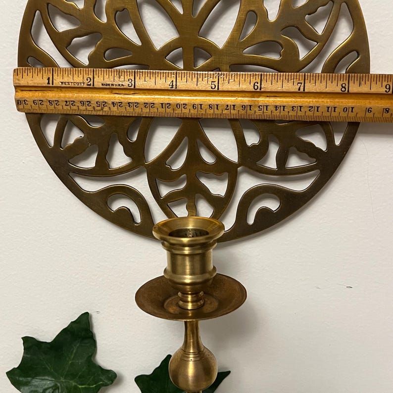 May include: A brass candle holder with a decorative, circular filigree design. The candle holder is approximately 9 inches in diameter. A wooden ruler is placed across the top of the candle holder to measure its size.
