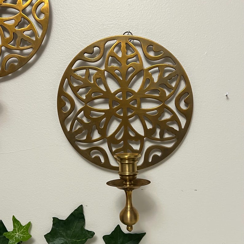 May include: Gold-colored metal wall sconce with a circular, openwork design. The sconce features a decorative floral pattern and a candle holder. The sconce is mounted on a white wall, with green leaves in the foreground.