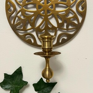 May include: A brass candle holder with a decorative, circular backplate featuring an openwork design. The candle holder has a small, round base and a cup-shaped top to hold a candle. Green leaves are in the foreground.
