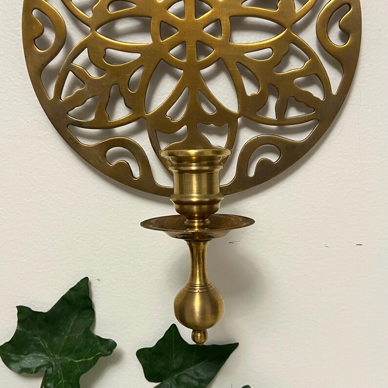 May include: A brass candle holder with a decorative, circular backplate featuring an openwork design. The candle holder has a small, round base and a cup-shaped top to hold a candle. Green leaves are in the foreground.