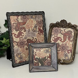 May include: Three decorative picture frames with a paisley pattern. The frames are in various shapes and sizes, with ornate detailing and a mix of colors including black, gold, and gray. The paisley design features shades of red, brown, and cream.