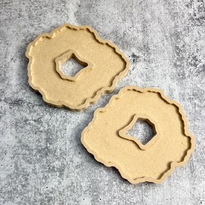 May include: Two light brown, agate-shaped coasters with a square cutout in the center. The coasters have a textured surface and a wavy edge. They are displayed on a gray, speckled surface.