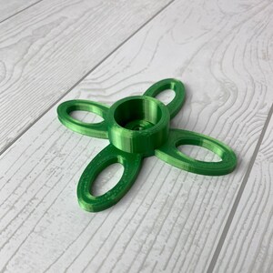 Puede incluir: Un portavelas verde impreso en 3D con cuatro brazos en forma de bucles. El portavelas tiene una abertura circular central para una vela.