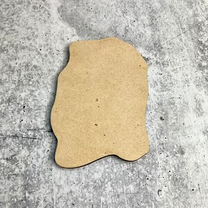 May include: A light brown, irregularly shaped wooden cutout, possibly for crafting or decoration. The cutout has a smooth surface and is set against a mottled gray background.