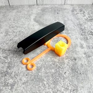 May include: A black, rectangular object and an orange and yellow tool are displayed on a gray surface. The tool has a circular handle and a square, yellow block. The black object is rectangular and has a curved top.