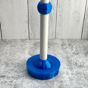 May include: A blue and white stand with a round base and a central pole. The base has a textured surface, and the pole is held in place by a blue ring. The stand is designed to hold an object.