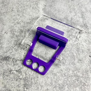 May include: A purple 3D printed phone stand with three holes for holding a phone. The stand is on a clear acrylic base.