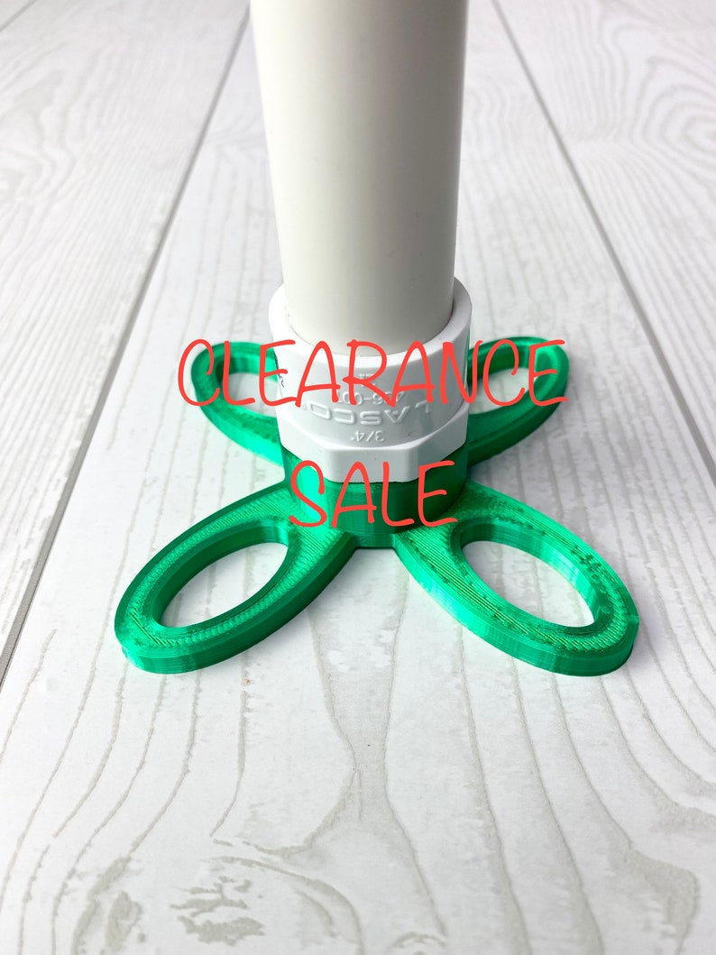 Puede incluir: Base de pl&aacute;stico verde con una conexi&oacute;n de tubo de PVC blanco. La base tiene cuatro brazos que forman una forma de tr&eacute;bol. El texto "CLEARANCE SALE" est&aacute; impreso en rojo en la base.