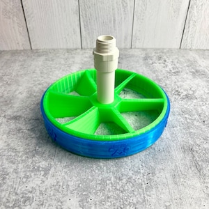 May include: A 3D-printed, multi-colored object featuring a circular base with a green interior and a blue rim. A white, cylindrical component extends upwards from the center. The blue rim has a paw print design. The object appears to be a pet-related product.