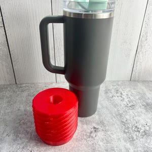 May include: A gray stainless steel travel mug with a lid and a handle. The mug is sitting on a white surface next to a red plastic object with a threaded design.