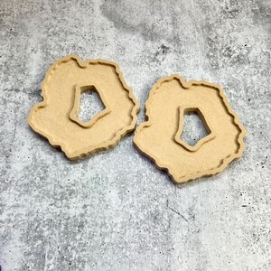 May include: Two light brown, wooden coasters with a unique, irregular shape and a central cutout. The coasters have a natural, unfinished appearance, suggesting a rustic or handmade aesthetic. They are placed on a textured, gray surface.