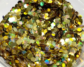 May include: A close-up of a clear plastic container filled with iridescent gold glitter. The glitter is hexagonal and has a variety of sizes.