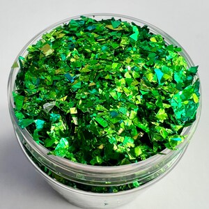 May include: A clear plastic container filled with vibrant green glitter flakes. The glitter has a reflective, iridescent quality, catching the light and creating a shimmering effect. The flakes are irregularly shaped, adding to the visual interest.