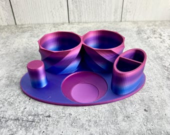 May include: A 3D printed desk organizer with a swirling gradient of pink, purple, and blue. The organizer has two large compartments, a small compartment, and a round base.