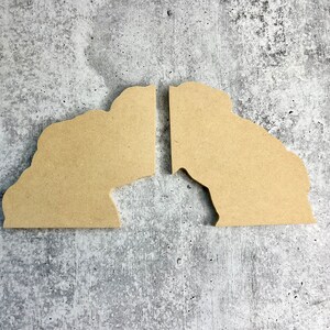 May include: Two unfinished wooden shapes, each resembling a person's head, are placed side by side on a gray textured surface. The shapes are facing each other, with a small gap between them.