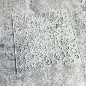 May include: A clear, square stencil featuring a repeating pattern of skull designs. The skulls are in various sizes and orientations, creating a textured effect. The stencil is made of a transparent material and is suitable for crafting projects.