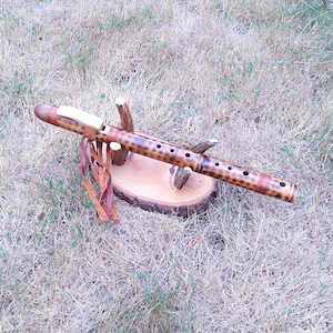 May include: A wooden flute with a spotted pattern, displayed on a small wooden stand. The flute is brown and tan, with leather detailing. The stand is a cross-section of a tree branch. The background is dry grass.