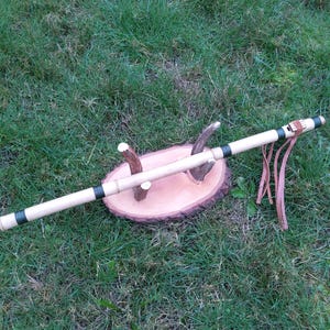May include: A handcrafted wooden flute with black accents and leather details rests on a wooden stand. The flute is light-colored with dark bands and leather straps. The stand is a cross-section of a tree trunk.