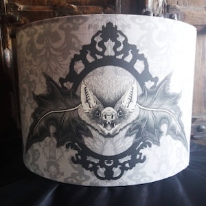 Puede incluir: Una pantalla de lámpara blanca con una ilustración en blanco y negro de un murciélago en un marco decorativo. El murciélago tiene orejas grandes y un hocico puntiagudo. El fondo es un patrón damasco gris y blanco.