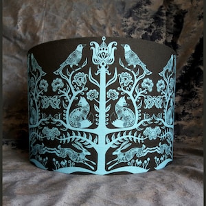 Puede incluir: Una pantalla de lámpara negra con un diseño azul claro de un árbol estilizado con pájaros, zorros, mariposas y liebres. El diseño recuerda al arte popular.