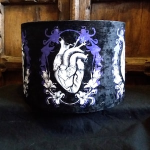 Puede incluir: Un tambor de terciopelo negro con un corazón anatómico blanco y diseños florales morados. El tambor está decorado con un patrón repetido de corazón y flores.