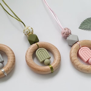 Peut inclure: Trois anneaux de dentition en bois pour bébé avec des accents en silicone colorés. Chaque anneau présente une combinaison de couleurs différente : gris, vert olive et rose. Les anneaux sont reliés à un cordon avec des perles assorties. Des fleurs décoratives sont en arrière-plan.