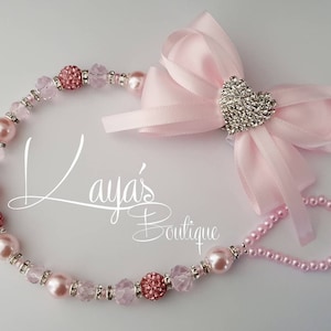 May include: Pink and clear beaded dummy clip with a pink satin bow and a rhinestone heart. The clip is attached to a pink satin ribbon.