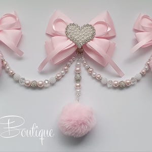 May include: Pale pink bow hair accessories with pearl and rhinestone details. Each bow has a fluffy pink pom-pom and is linked by a beaded chain. The central bow features a heart-shaped rhinestone embellishment.