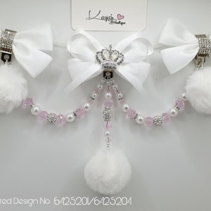 May include: Three white satin bows with rhinestone accents and white pom-poms attached to a pink and white beaded chain.  The centre bow has a silver crown with rhinestones.  Registered Design No 6425201/6425204