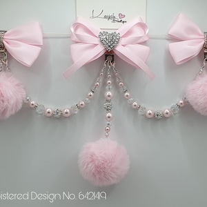May include: Three pink satin bows with silver clips and rhinestones, connected by a silver chain with pink and clear beads and three pink fluffy pom-poms. Registered Design No. 6421419