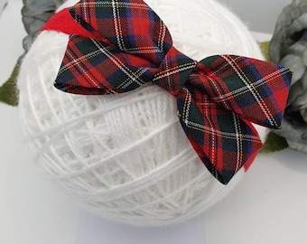 Puede incluir: Una pajarita a cuadros rojos, verdes y azul marino está centrada en una bola de hilo blanco. La pajarita es de tela y tiene una forma clásica. El fondo incluye una superficie blanca y una etiqueta con el texto "Kaya's Boutique".