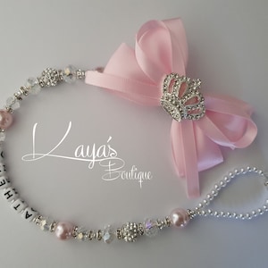 May include: A pink and silver dummy clip with a crown charm and a pink bow. The clip is personalised with the name "ATHENA".