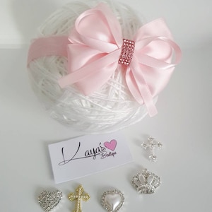 May include: A pink satin bow with rhinestones on a white yarn ball.  There are five decorative embellishments: a silver heart, a gold cross, a silver crown, a silver heart with a pearl center, and a gold cross with pearls.  The card reads "Laya's Boutique".