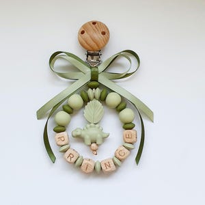 May include: A baby's dummy clip with a wooden clip and a green ribbon bow. The clip features a ring of green beads and wooden letter blocks spelling "PRINCE", with a green dinosaur charm in the centre.