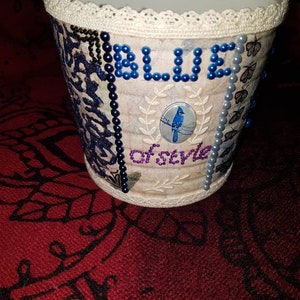 Peut inclure: Un pot en céramique blanc avec un motif d'oiseau bleu et le mot "BLUE" écrit en perles bleues. Le pot est orné d'une bordure en dentelle autour du haut et est décoré de divers embellissements, notamment des perles, des papillons et des motifs floraux.