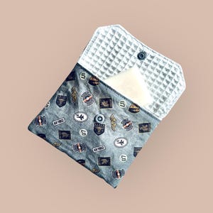 Puede incluir: Una bolsa de tela gris con un forro blanco de textura de gofre. La bolsa está decorada con un patrón repetido de parches de inspiración militar, incluyendo las palabras "Air Force" y "07". La bolsa está abierta y un objeto rectangular blanco es parcialmente visible en el interior.