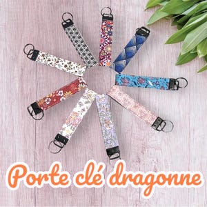Peut inclure: Un ensemble de porte-clés colorés à motifs avec des anneaux métalliques noirs. Les porte-clés sont disposés en cercle sur une surface en bois. Le texte "Porte clé dragonne" est visible en bas de l'image.